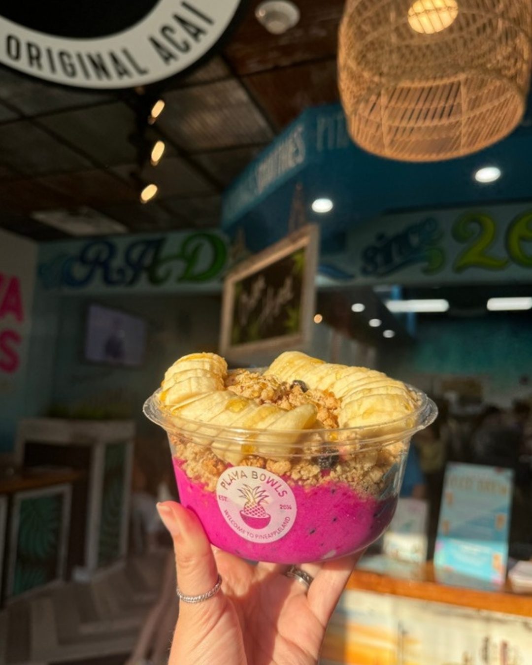 A pitaya bowl topped with banana and granola, held up inside a Playa Bowls shop