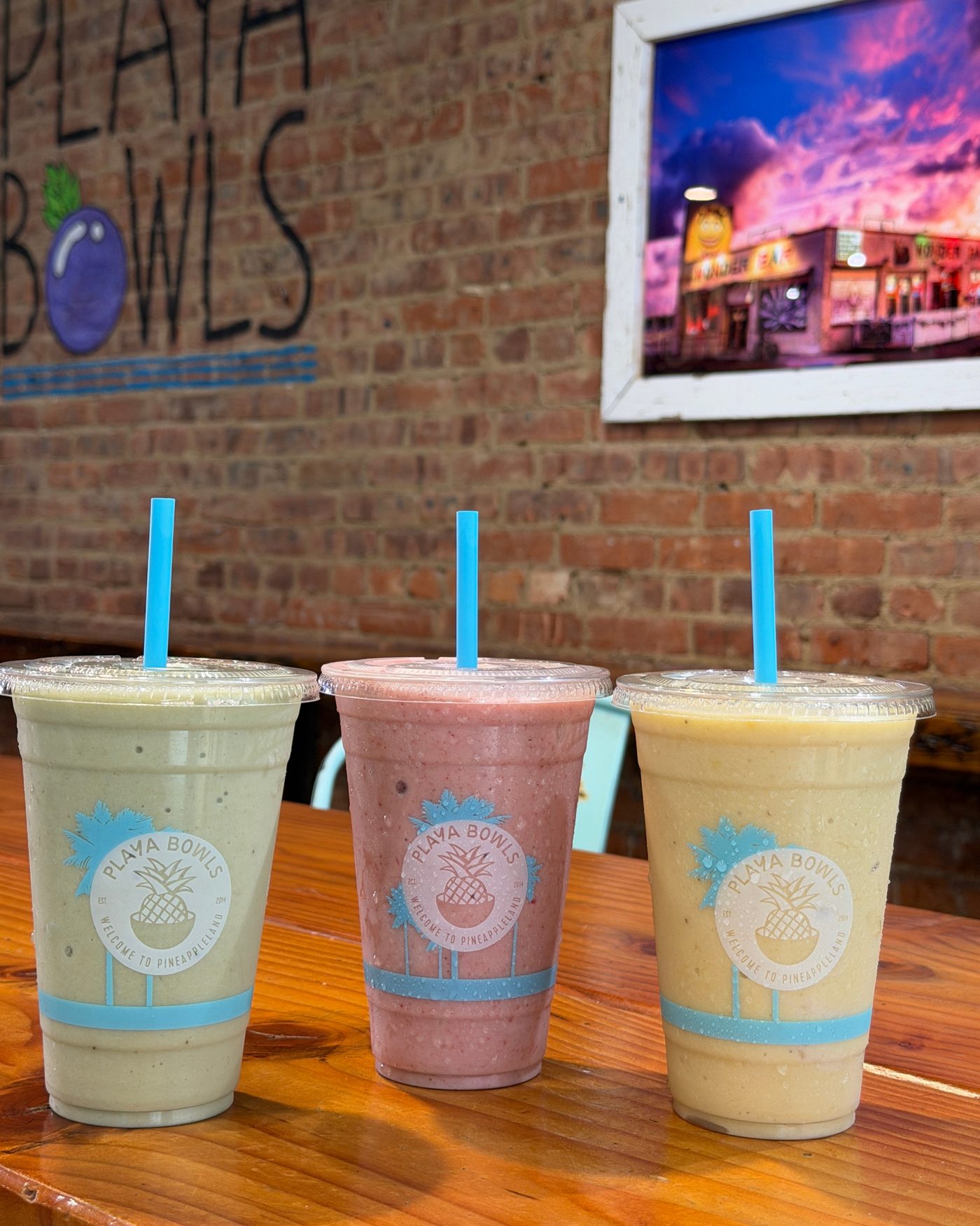 Three Playa Bowls smoothies in branded cups on a wooden table